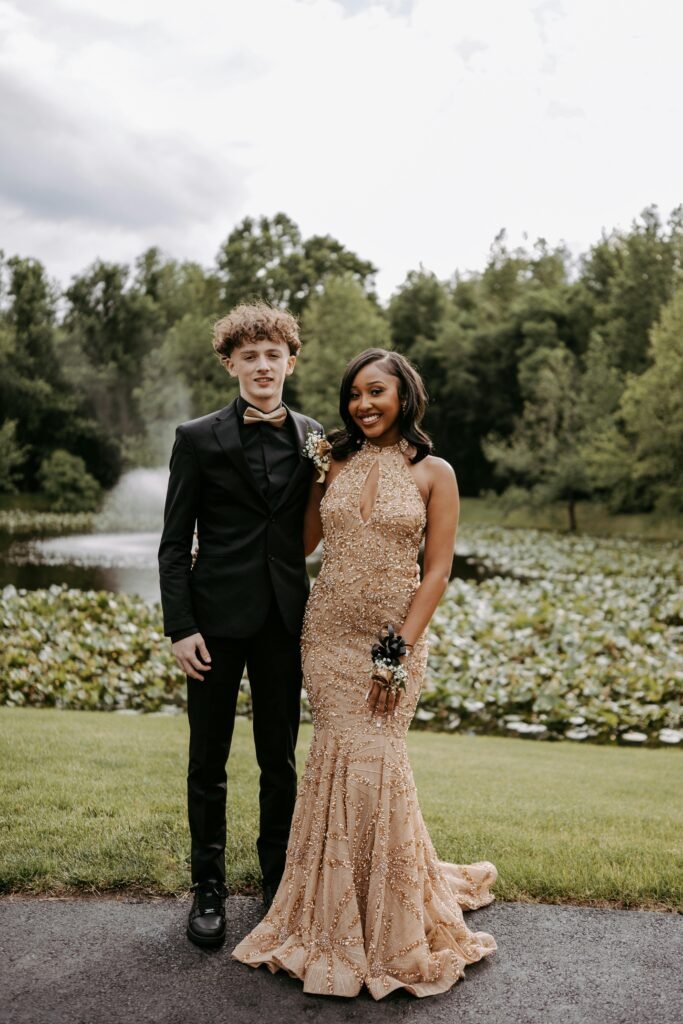 A stylish prom couple posing by a beautiful pond surrounded by lush greenery.